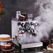 La Pavoni Cellini Classic Traditional Model espresso machine in sleek stainless steel, steaming on a countertop with coffee accessories, ceramic cups, and desserts. Professional-grade equipment shown in atmospheric setting with dramatic steam and modern styling.