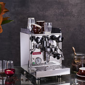 La Pavoni Cellini Classic Traditional Model espresso machine in polished stainless steel with dual gauges, steam wand, and portafilter displayed on counter with coffee cups and beans, set against dark textured background with decorative plant accent